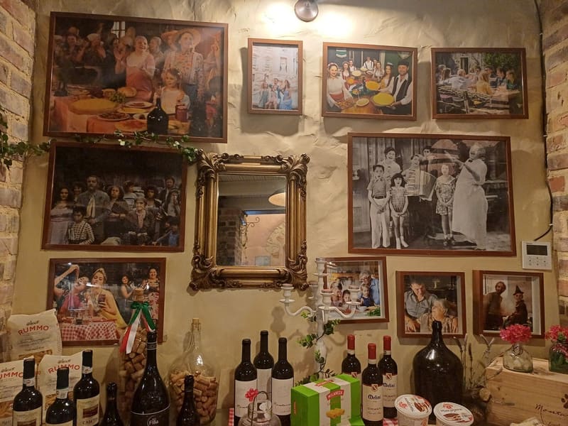Parede de fotografias antigas de famílias à mesa, com um espelho dourado, sobre garrafas de vinho