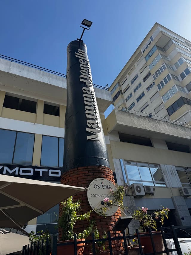 Garrafa gigante com o nome Mamma Donella sobre uma base de tijolo, com o disco «Osteria, cozinha italiana»