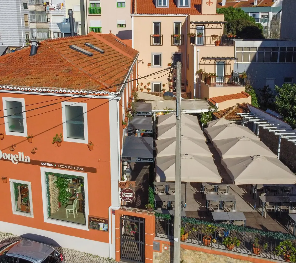 A casa de Massamá vista de cima: a fachada terracota com o letreiro e a placa redonda, e ao lado a esplanada com chapéus de sol